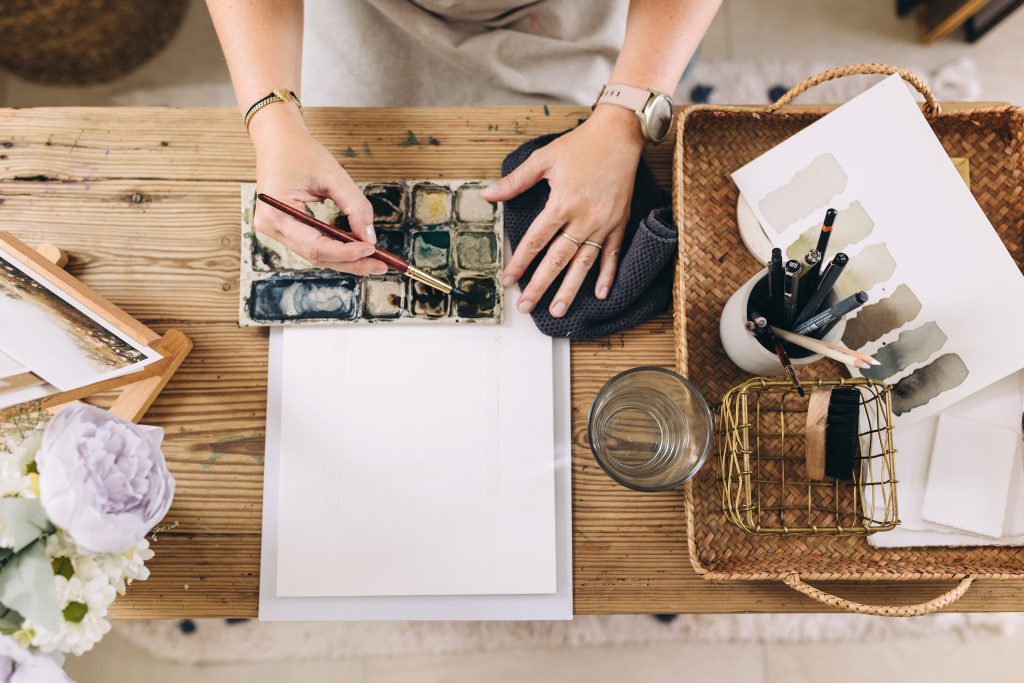 Creativity is the main theme here. This image contains art supplies on a table. A woman is sitting at the table and her hands are shown using a paintbrush in watercolors and a blank canvas in front of her. 