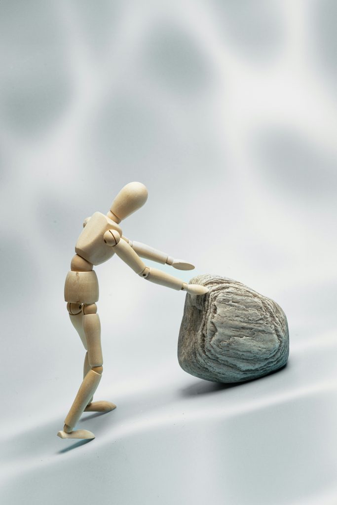 this is an image of a male wooden puppet figure pushing a large boulder up a hill